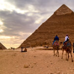 Three Tourists Are Riding Camel And Moving To The Pyramid Three Tourists Are Riding Camel And Moving To The Pyramid