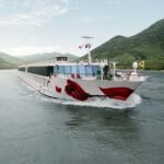 Ein elegantes, weißes Flusskreuzfahrtschiff der AROSA Flotte mit einem auffälligen roten Rosenmotiv an der Bugseite, das sich auf einem breiten Fluss zwischen sanften, bewaldeten Hügeln bewegt. Der Himmel ist leicht bewölkt und das Wasser zeigt leichte Wellen durch die Fahrt des Schiffes. Ein elegantes, weißes Flusskreuzfahrtschiff der AROSA Flotte mit einem auffälligen roten Rosenmotiv an der Bugseite, das sich auf einem breiten Fluss zwischen sanften, bewaldeten Hügeln bewegt. Der Himmel ist leicht bewölkt und das Wasser zeigt leichte Wellen durch die Fahrt des Schiffes.
